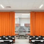 Curtain Walls for Blocking Off Cafeteria Equipment During Non-Use Hours, orange vinyl curtains on a ceiling track closing the kitchen behind dining tables