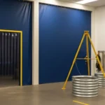 Blue vinyl curtain wall with a clear strip-door entrance used to separate an indoor confined-space practice zone with tripods and metal entry drums