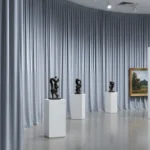 Gray museum exhibit divider curtains providing a soft backdrop for sculptures and framed landscape paintings