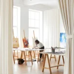 White floor-to-ceiling curtains dividing a sunlit art studio with easels