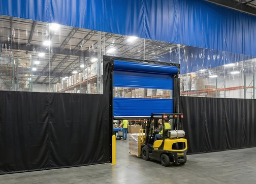 Forklift driving through a blue powered roll-up door set in a black and clear vinyl warehouse curtain wall